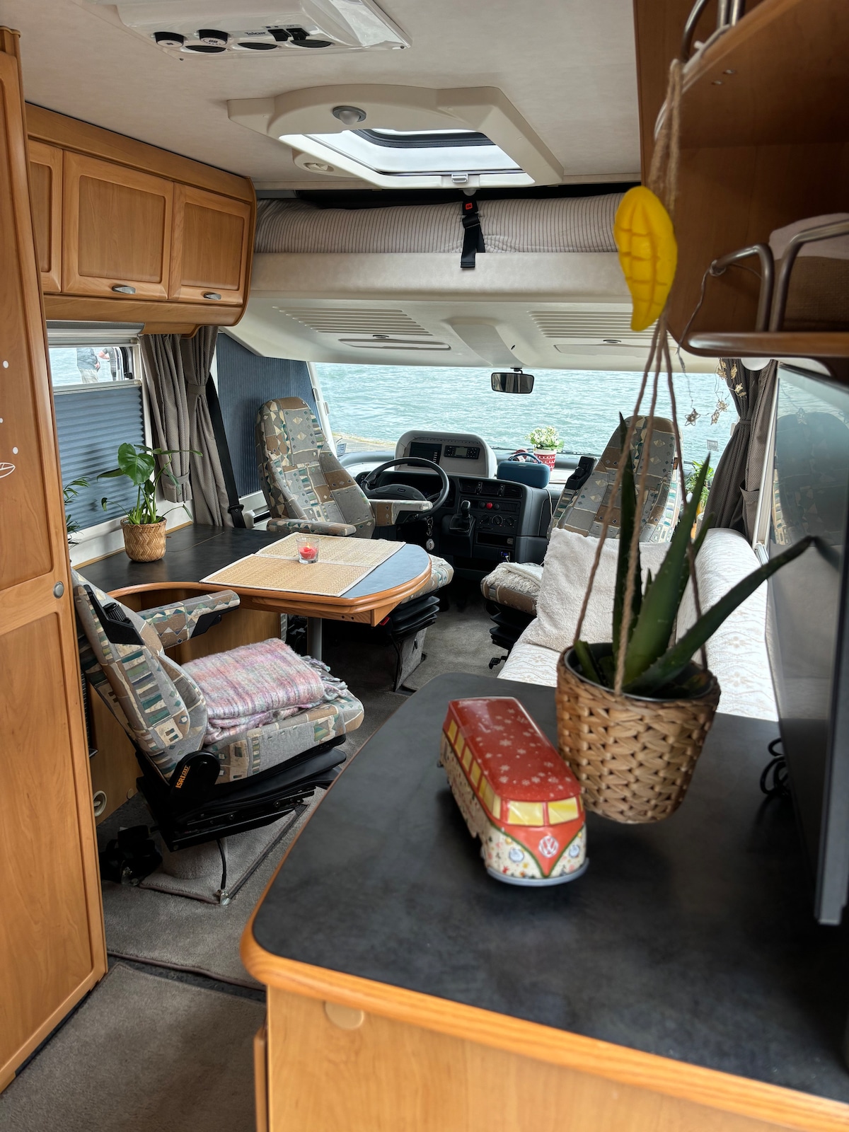 A cozy interior of a camper van features wooden cabinetry and comfortable seating with patterned upholstery. A small dining area is present with a folding table and potted plants. Natural light enters through a skylight, enhancing the spacious feel of the living area.