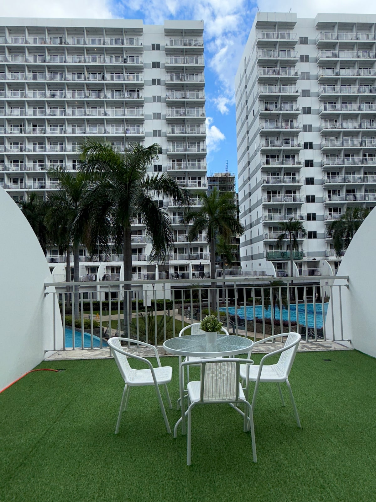 A balcony area is presented, featuring a glass-top table accompanied by four white chairs. Lush green artificial turf covers the floor. In the background, two modern buildings with multiple balconies and palm trees are visible, alongside two swimming pools reflecting the sky.