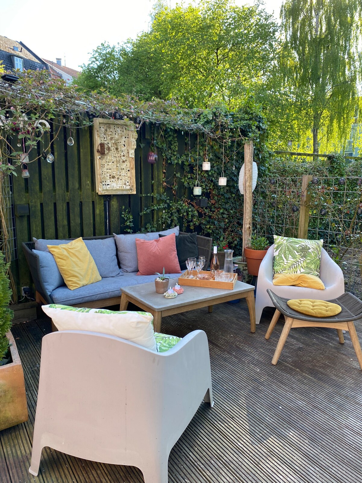 A serene outdoor lounge area is depicted, featuring a cozy couch adorned with colorful cushions and a wooden coffee table. Surrounding greenery provides a natural backdrop, while additional seating is offered by two white chairs with patterned pillows, creating an inviting space for relaxation.