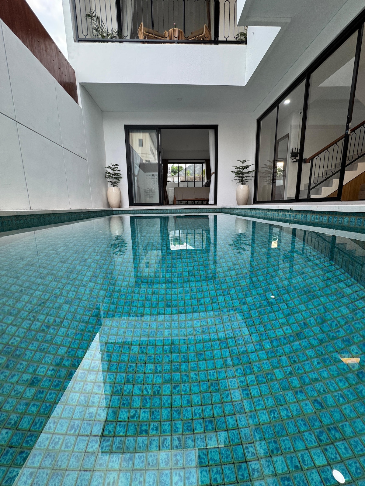 A swimming pool reflects the vibrant blue tiles and the white walls surrounding it, creating a serene atmosphere. Large glass doors, partially visible, open towards a living space inside the villa, enhancing connectivity between indoor and outdoor areas.