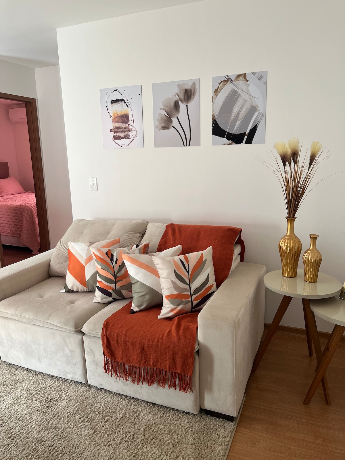 A neutral-toned couch is adorned with patterned cushions and a vibrant throw. Above, framed artworks add visual interest to the wall. A small table is accompanied by two decorative vases, contributing to the inviting ambiance of the cozy living space.