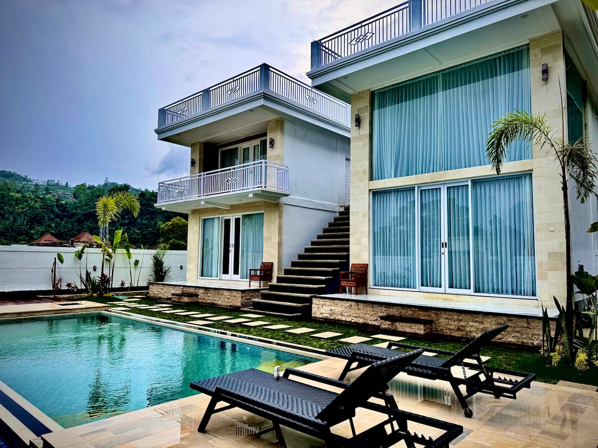 The villa features a contemporary design with large glass windows and balconies. A swimming pool is situated in front, complemented by lounge chairs. Lush greenery surrounds the property, enhancing the outdoor space. A staircase leads to the upper level, providing access to additional living areas.