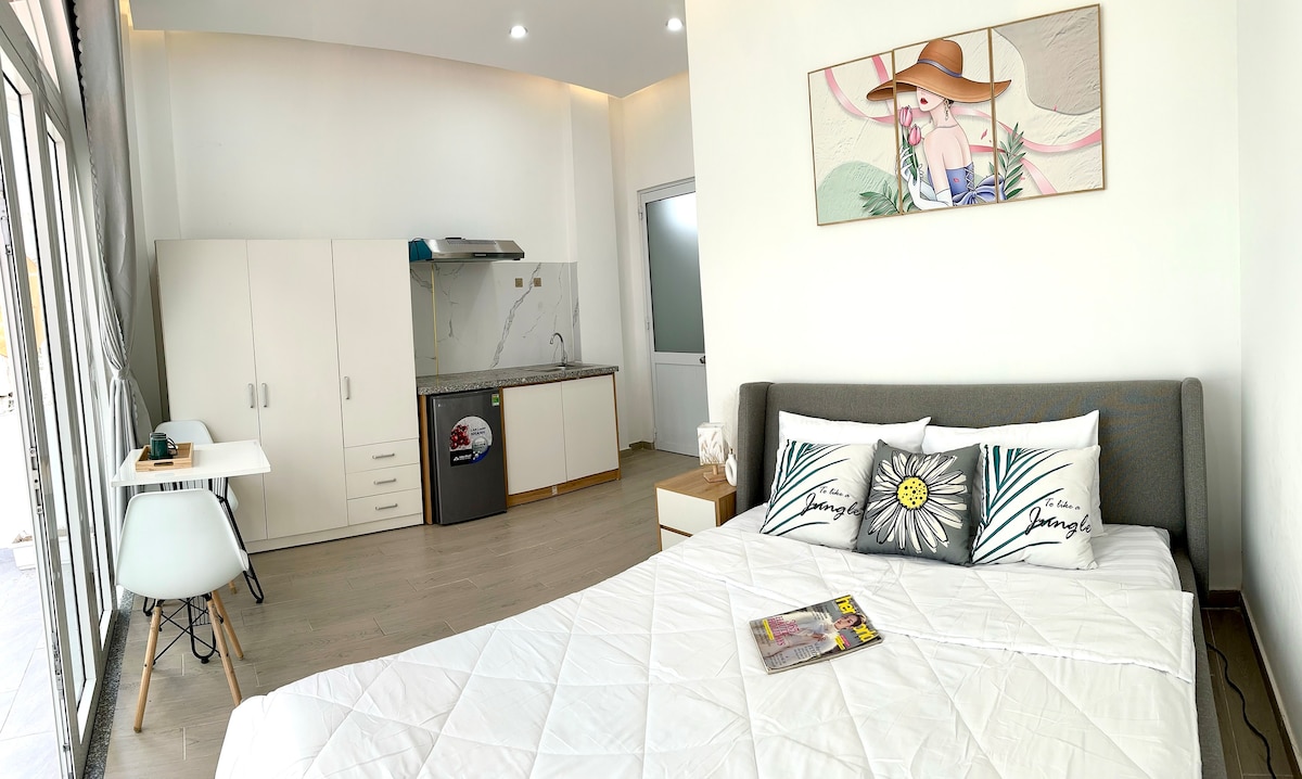 A well-lit studio apartment is presented, featuring a spacious bed adorned with decorative pillows. A small dining table and chairs are positioned nearby, while a kitchenette equipped with modern appliances is visible in the background. Natural light enters through expansive windows.