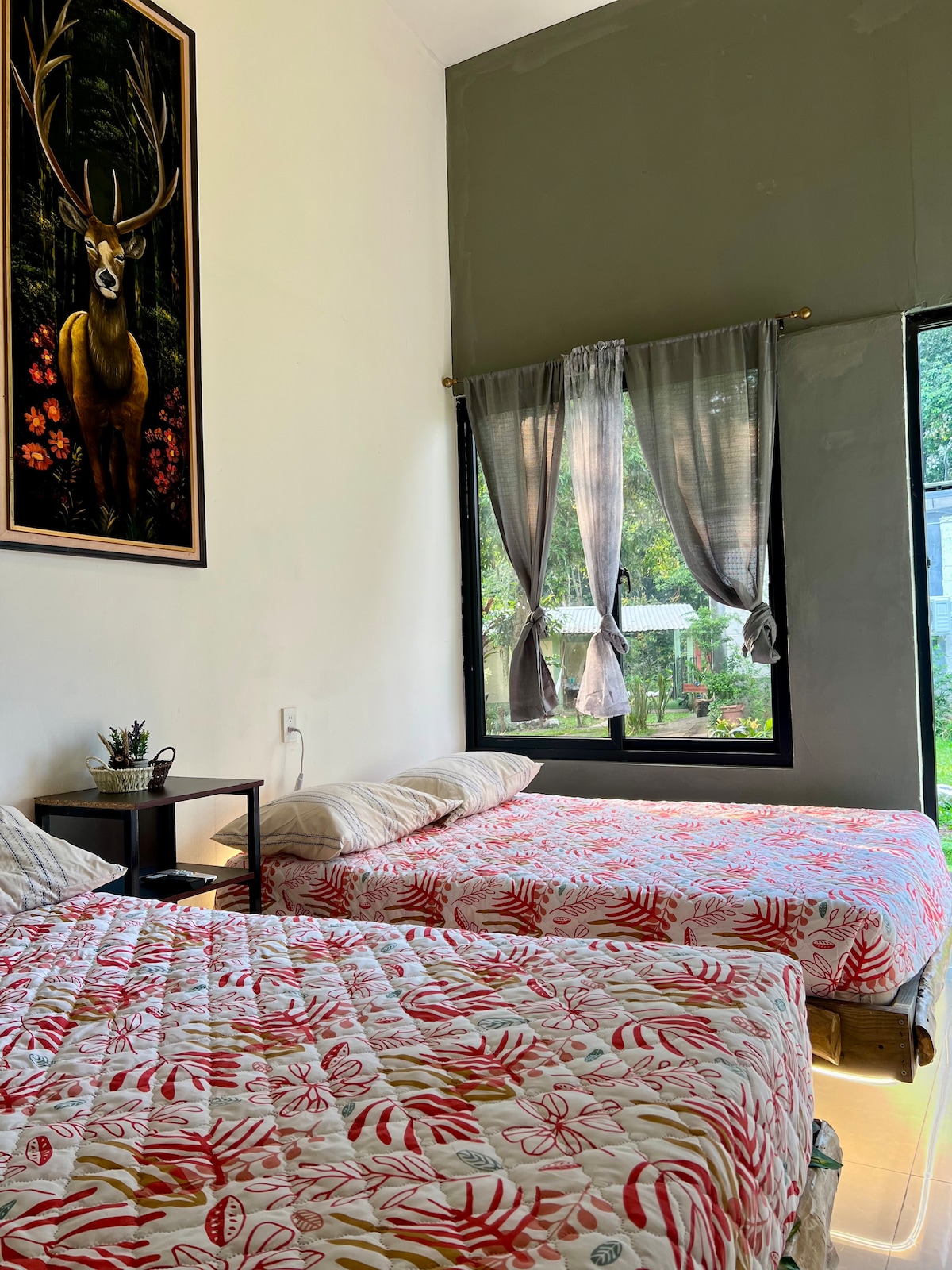 A guest room is featured with two spacious beds, adorned with floral patterned bedding. Natural light filters through a large window, which is framed by sheer curtains. A framed artwork of a deer adorns the wall, complementing the serene atmosphere.