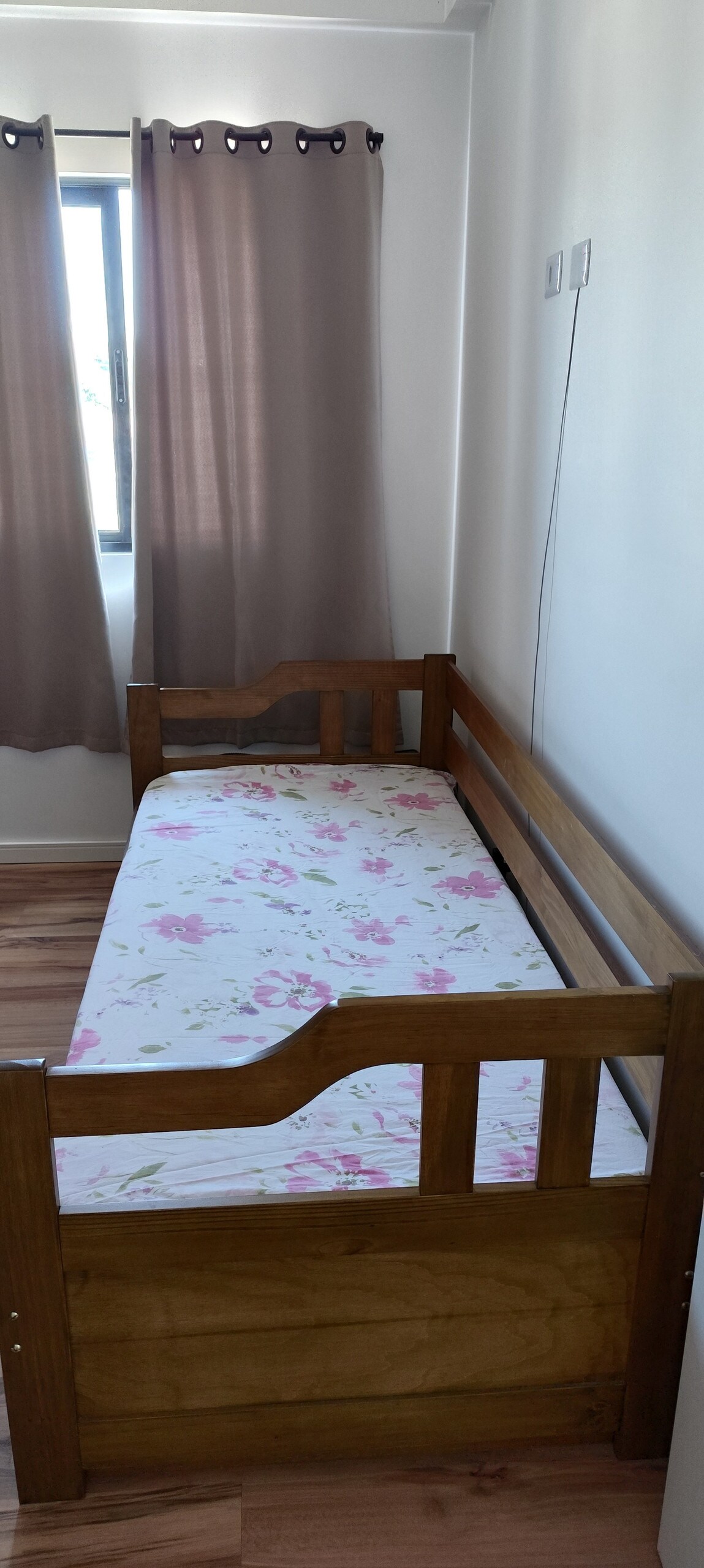 A single bed with a floral-patterned mattress is positioned against a light-colored wall. Natural light enters the room through a nearby window, which is covered by sheer curtains that diffuse the sunlight. Wooden accents provide a simple and functional design.