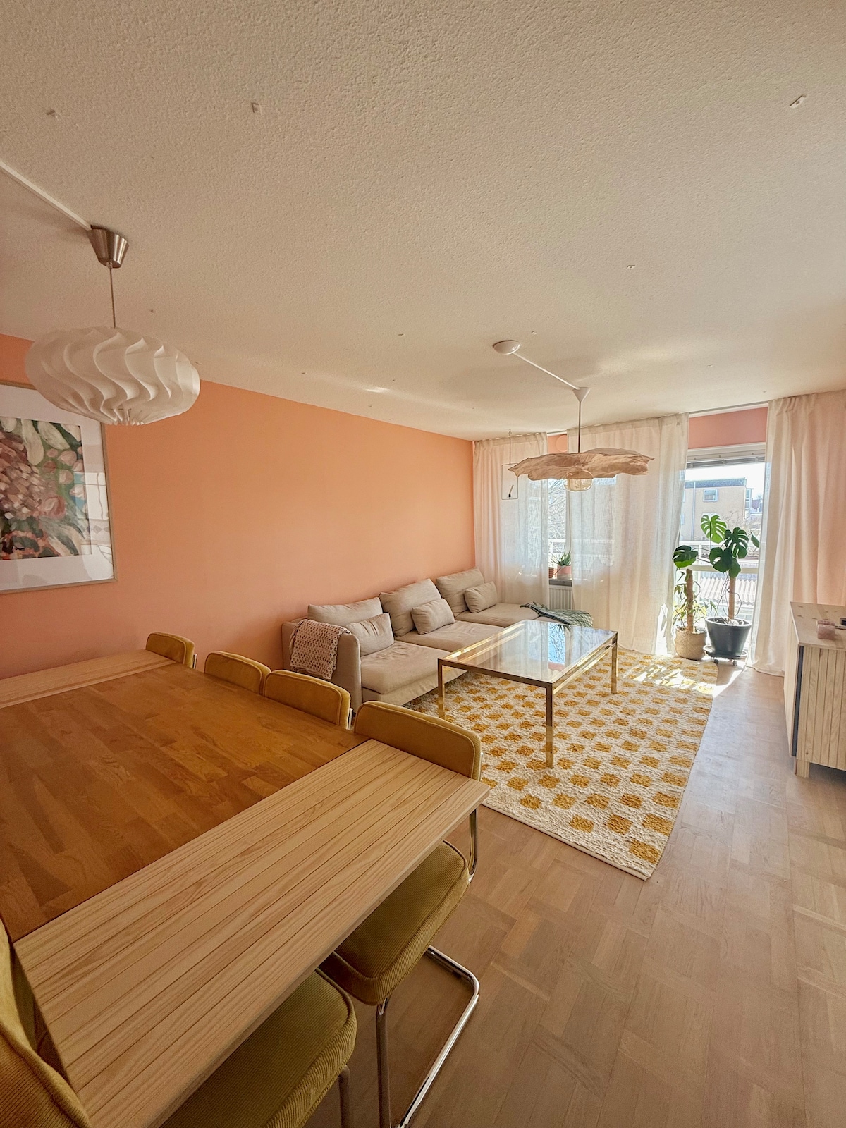 A light-filled living area features a cozy sofa and a glass coffee table on a patterned rug. A wooden dining table with upholstered chairs is visible, complemented by soft furnishings and large windows that allow for ample natural light.