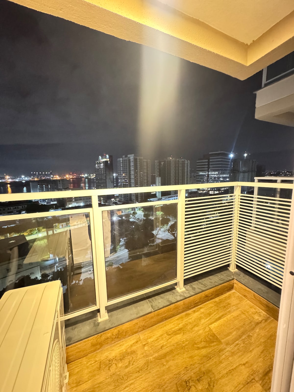 A spacious balcony presents a view of the city skyline at night, illuminated by numerous buildings and lights. The railing features horizontal slats, providing a modern aesthetic. A cooling unit is positioned to the side, and the floor is laid with wooden tiles.