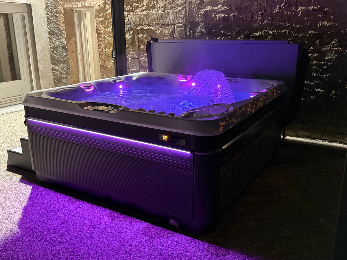 A luxurious Caldera Utopia spa is positioned on a textured outdoor surface, featuring soft, ambient purple lighting. The spa is equipped with various jets and a water fountain, offering a relaxing space for up to five individuals.