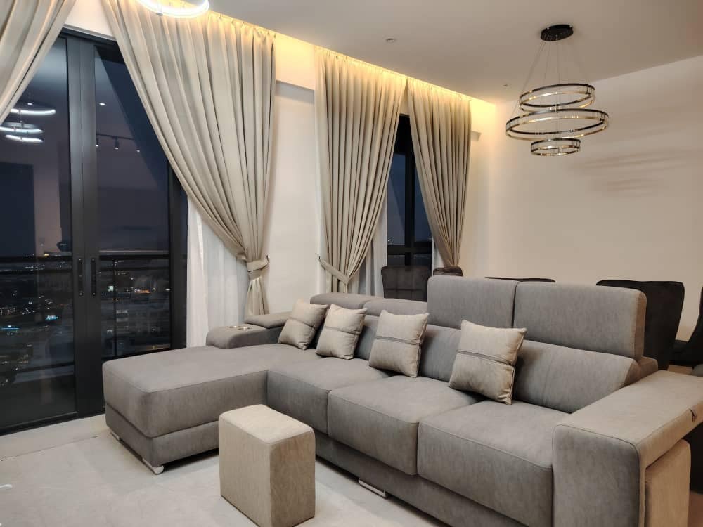 A modern living area is shown with a spacious gray sectional sofa adorned with several small pillows. Floor-to-ceiling windows allow views of the city at night. Elegant curtains frame the windows, and a pendant light fixture adds illumination.