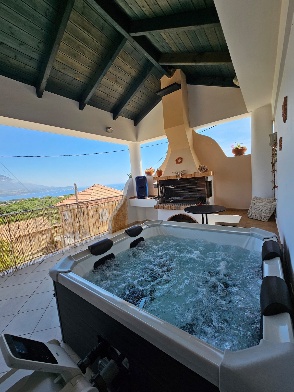 A spacious outdoor area features a hot tub and a fireplace under a vaulted ceiling, complemented by large windows that showcase sweeping views of mountains and sea. Potted plants add subtle greenery to the area.