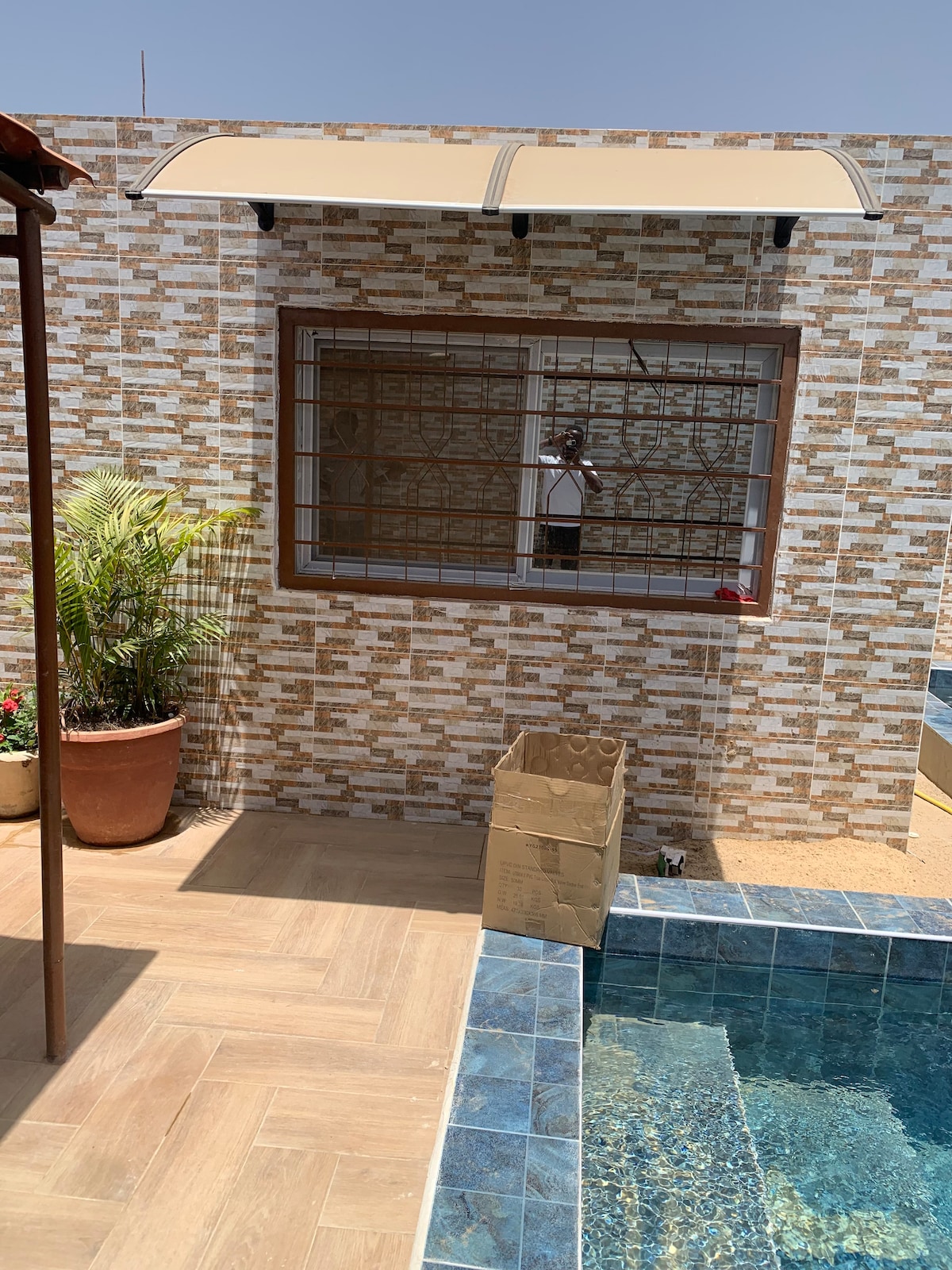 A small outdoor space is showcased, featuring a tiled pool reflecting sunlight. A decorative wall with a mosaic pattern serves as a backdrop. Potted plants add a touch of greenery, while a shaded area from an awning provides relief from the sun.