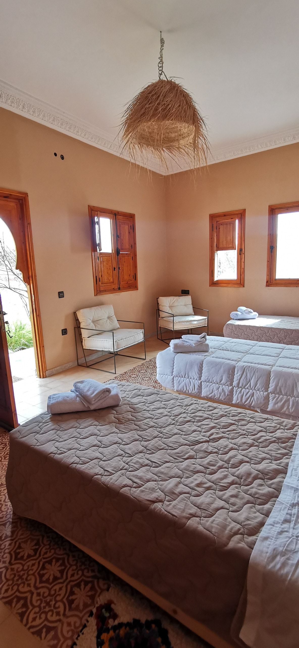 A spacious room features two neatly arranged beds with light-colored bedding. Natural light enters through multiple wooden windows, and a rattan light fixture hangs from the ceiling. A small seating area with two chairs is visible near a window, creating a bright and airy environment.