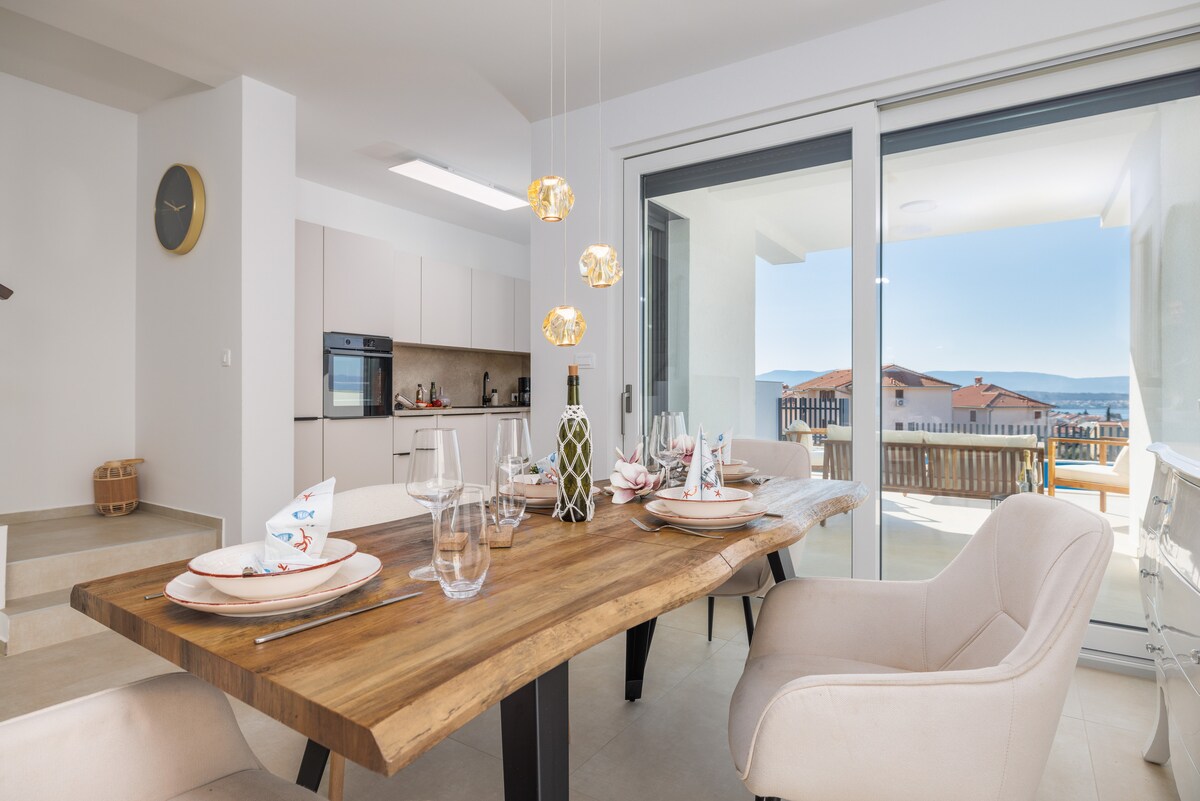 An open dining area features a wooden table set for six, complemented by modern chairs. Soft lighting from pendant lights adds a touch of elegance. Large sliding glass doors offer views of the outdoor terrace, creating a bright and airy atmosphere.
