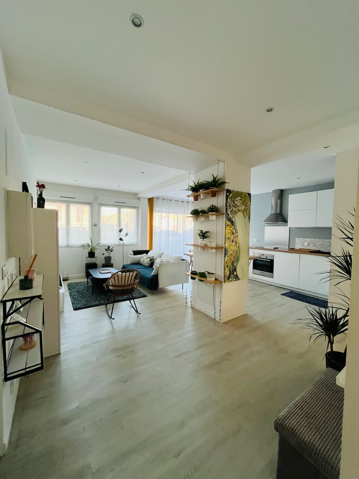 An open living space is observed, featuring a light-colored floor and bright walls. A cozy seating area is visible with a small coffee table. The kitchen area showcases modern appliances and cabinetry. Natural light filters through large windows, and green plants are arranged on shelves.