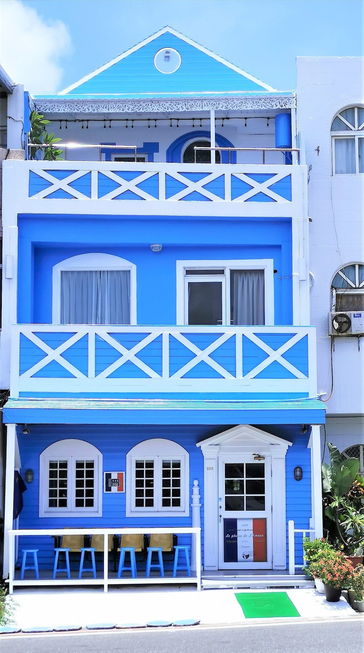 A charming blue building features a white-trimmed entrance with large windows, creating a welcoming facade. A spacious balcony with railings overlooks the street below. Outdoor seating is provided in front, complementing the vibrant color scheme of the structure.