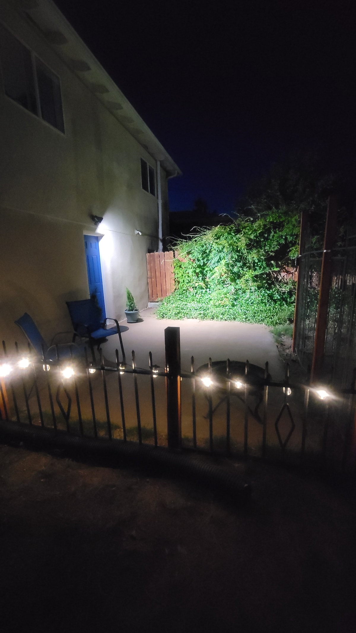 A well-lit outdoor area features a concrete patio enclosed by a decorative fence. Two blue chairs are positioned on the patio, with soft lighting created by string lights. A lush, green plant covers part of a nearby wooden fence, adding a touch of nature.