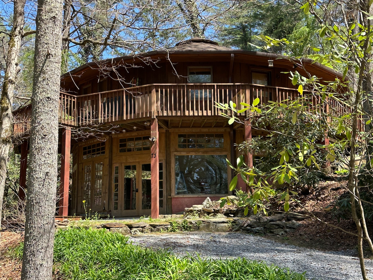 A roundhouse, constructed of natural wood, is situated among trees. Large windows allow ample light into the living spaces. A wrap-around deck extends from the front, and a gravel path leads to the entrance, bordered by lush greenery.