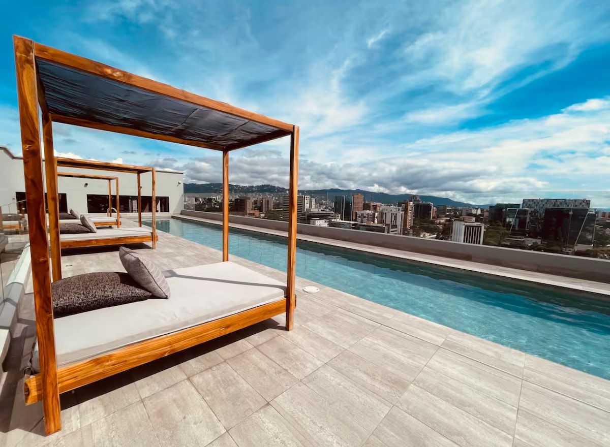 A rooftop infinity pool is flanked by wooden cabanas with padded seating. The serene water reflects the expansive sky overhead, providing panoramic views of the surrounding cityscape. The vibrant backdrop enhances the relaxing atmosphere of the space.