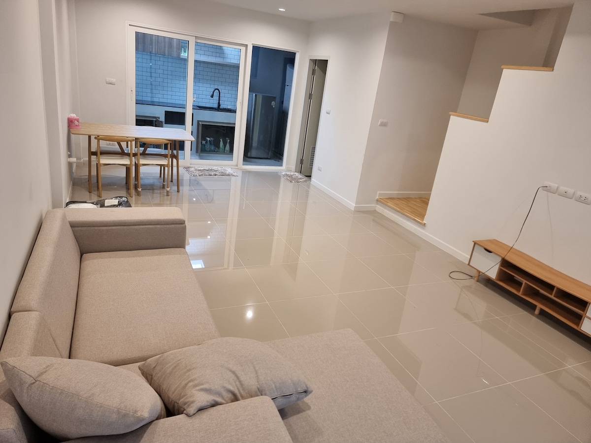 A spacious living area is shown with a light-colored sectional sofa positioned in the foreground. A wooden dining table is visible in the background, adjacent to large glass doors that lead to an outdoor space. Neutral tones and glossy flooring enhance the open atmosphere.