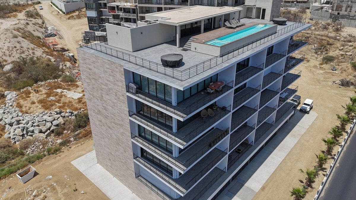 An aerial view of a five-story apartment building is presented, featuring a rooftop pool and spacious terrace. Balconies equipped with outdoor seating are visible on each floor. A surrounding landscape includes gravel and construction areas, indicating ongoing development in the locale.