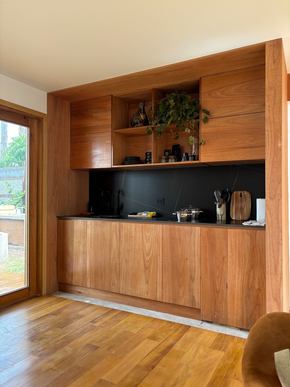 A modern kitchen area is highlighted by rich wooden cabinetry and a sleek black countertop. Natural light enters through a large window, enhancing the warm tones of the wood. Various kitchen utensils and decor items are neatly arranged on the counter, providing a functional and stylish space.