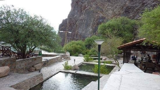 A serene outdoor area is depicted featuring natural rock formations in the background. The setting includes small, clear pools of water surrounded by lush green shrubs and trees. A shaded seating area is visible to the right, providing a space for relaxation.