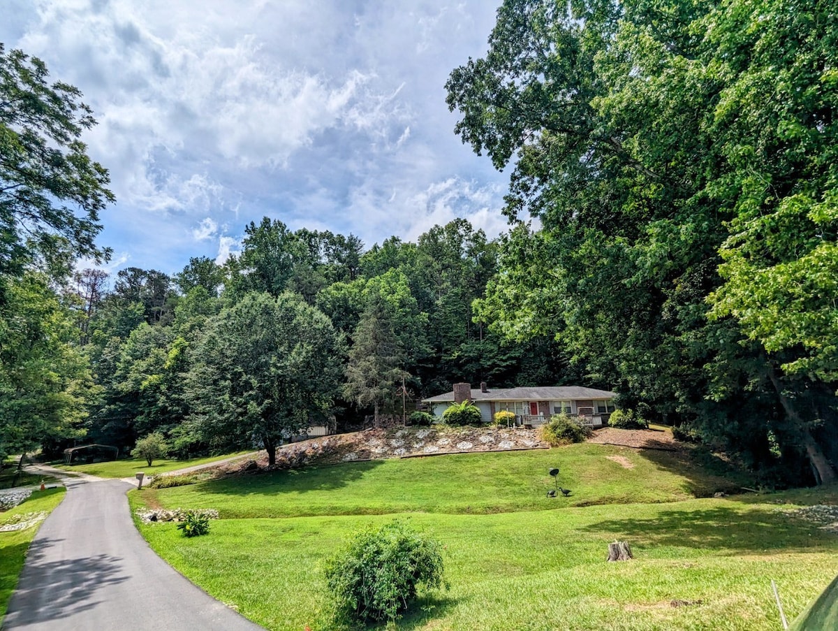 A spacious house is nestled among verdant trees and landscaped gardens, highlighting a serene environment. A winding driveway leads toward the home, framed by lush green grass and natural rock features. The sky above is partly cloudy, creating a peaceful backdrop.