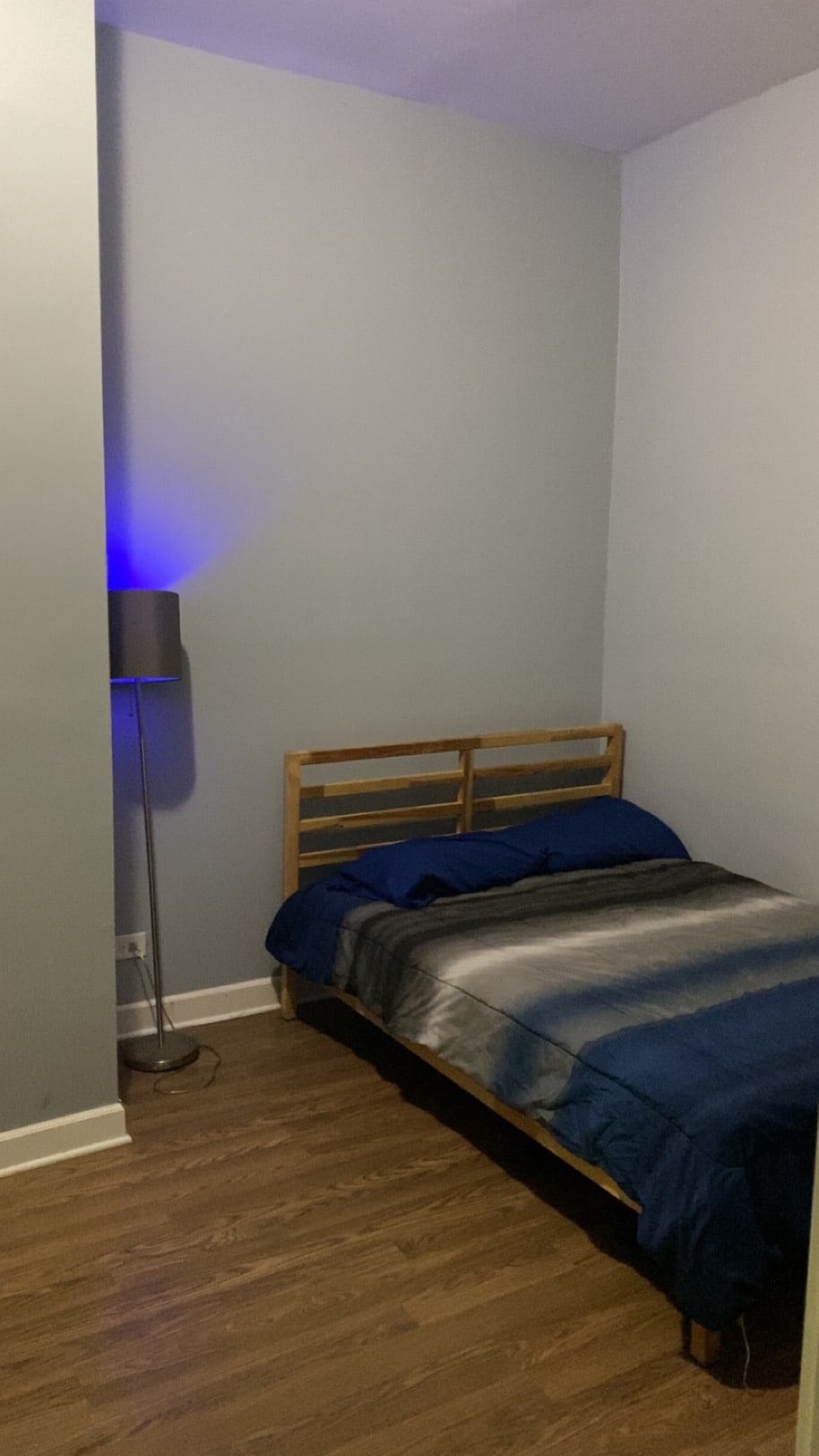 A cozy sleeping area is presented with a wooden bed frame featuring a blue and silver striped blanket. A modern floor lamp emits a soft blue light, adding a calming touch to the space. The walls are painted in a light shade, and the flooring is wooden.