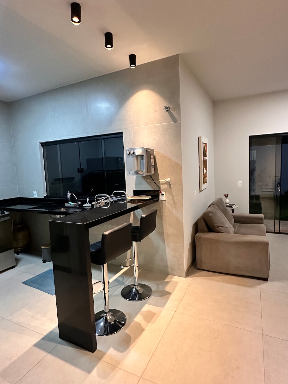 A modern kitchen area features sleek black countertops and a bar with two stylish stools. Natural light enters through a large window, illuminating the space. Adjacent to the kitchen, a cozy seating area is presented with a neutral-toned armchair, creating an inviting atmosphere.