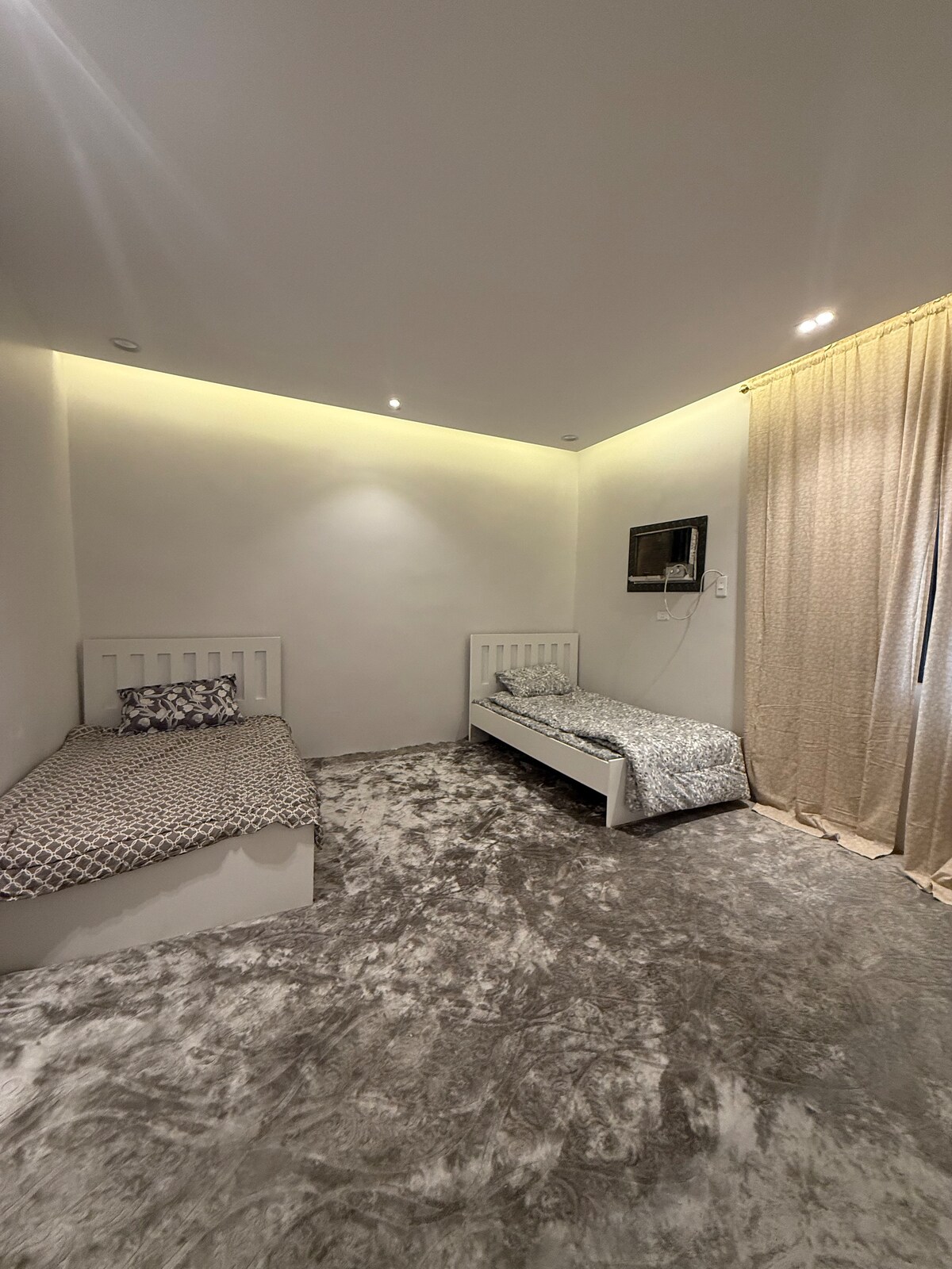 A well-lit bedroom features two spacious beds, each adorned with patterned bedding. A flat-screen TV is mounted on the wall. Soft, plush carpeting covers the floor, and natural light filters through a sheer curtain, creating a comfortable atmosphere.