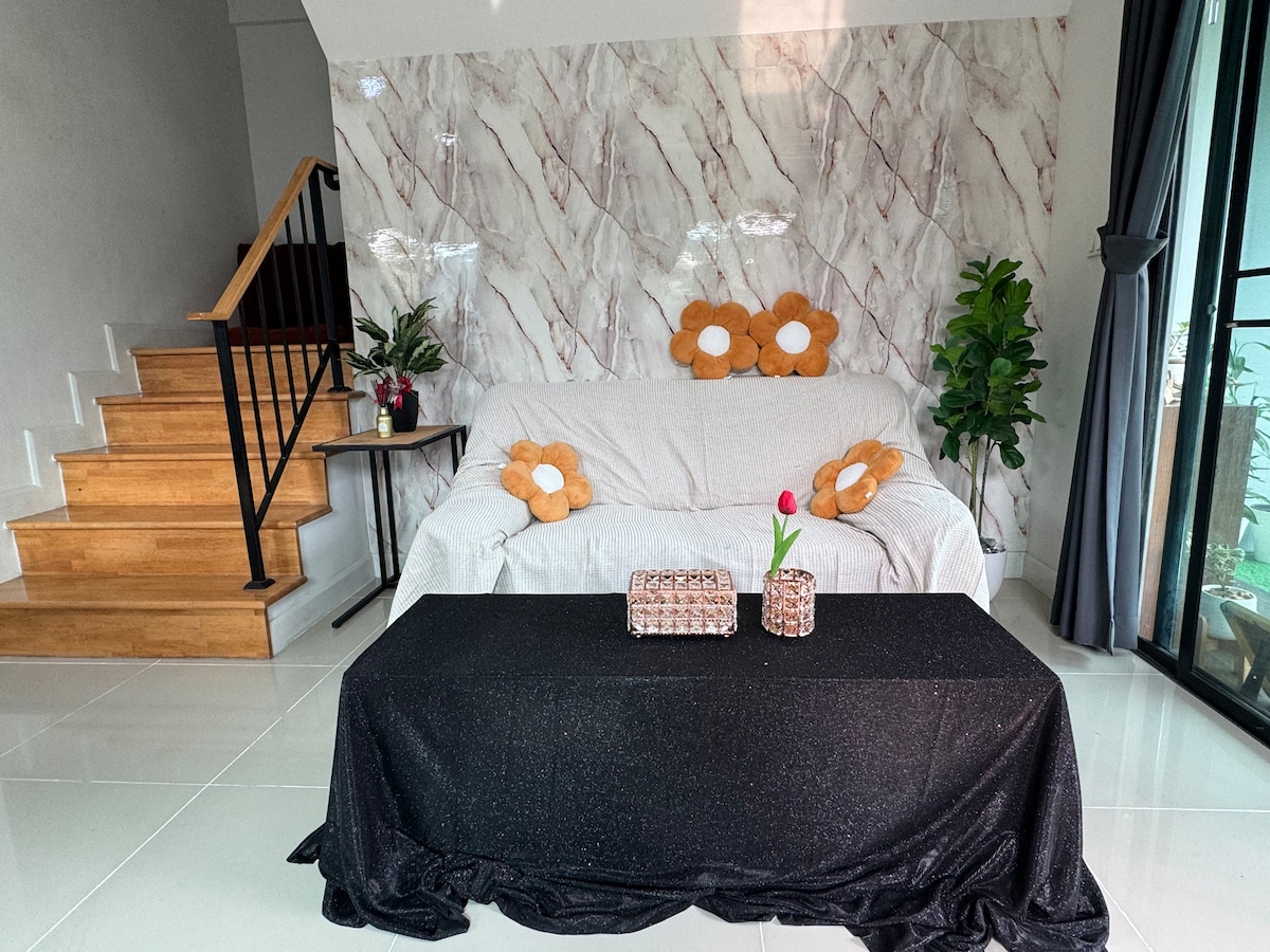 A living area features a light-colored sofa adorned with floral-patterned cushions. A dark table is positioned in front, complemented by decorative items. Sunlight filters through large glass doors, illuminating the space, while a potted plant adds a touch of greenery to the room.
