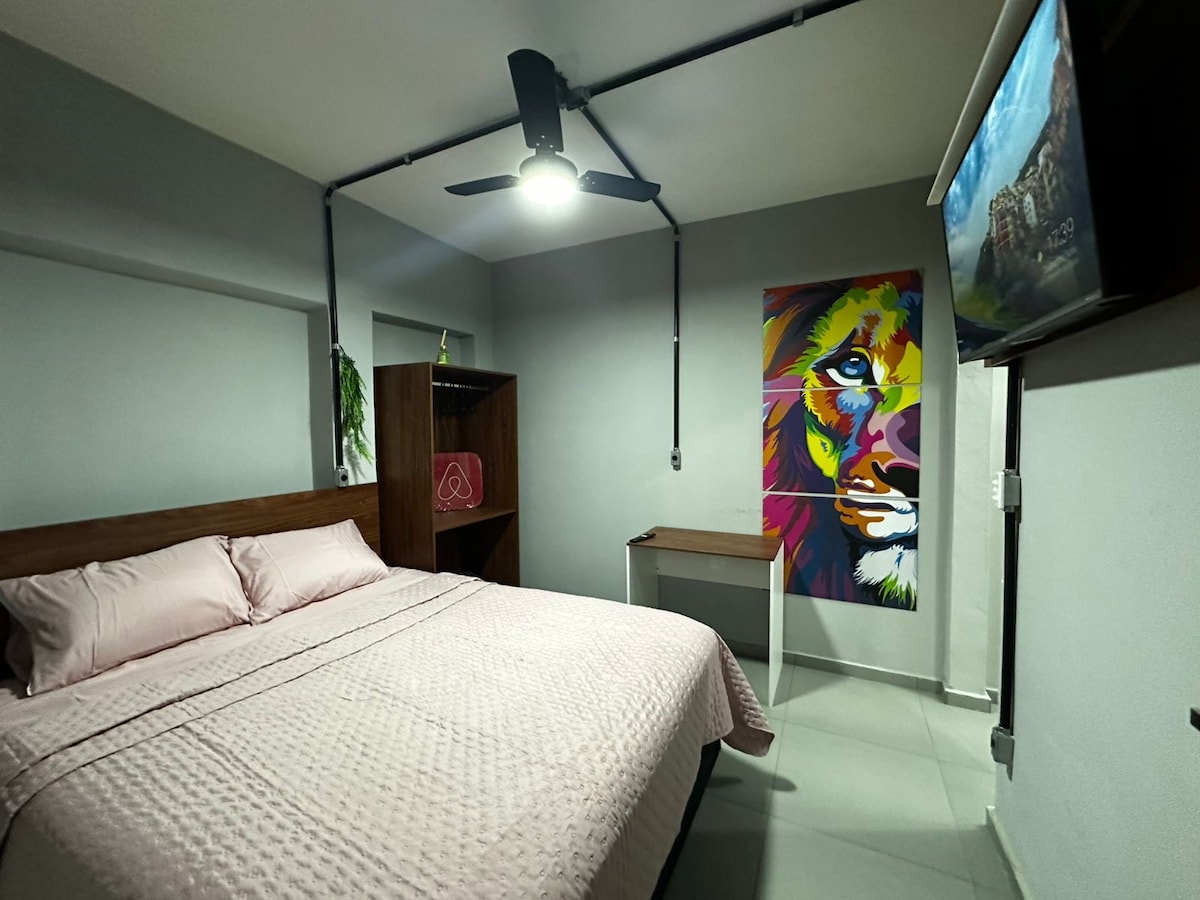 A well-appointed bedroom is featured with a spacious bed covered in light pink bedding. A vibrant lion mural adds color to the wall. A small desk is situated near a wall-mounted television, with a ceiling fan providing a cooling breeze.