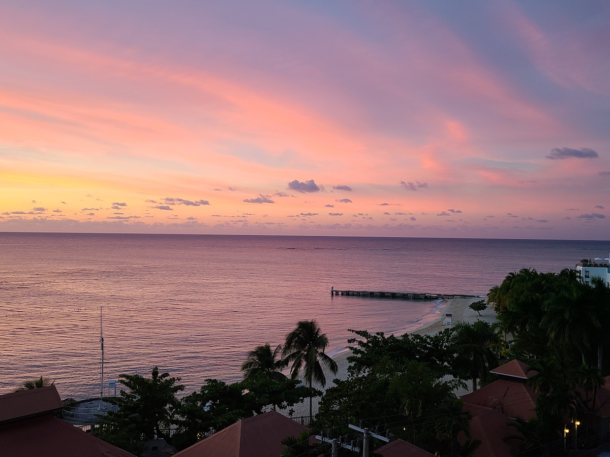 A serene sunset view showcases soft pink and purple hues across the sky as the ocean reflects the colors. Palm trees frame the scene, while a distant pier extends into the calm waters. The tranquil beach is illuminated by the fading light.