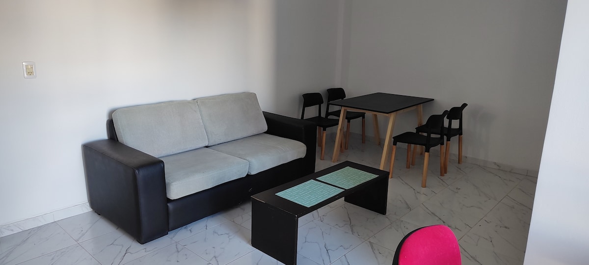 A modern living area features a two-tone sofa positioned beside a low coffee table. A rectangular dining table is accompanied by four chairs, creating a functional space for relaxation and dining. The light-colored flooring adds to the overall bright and airy ambiance.