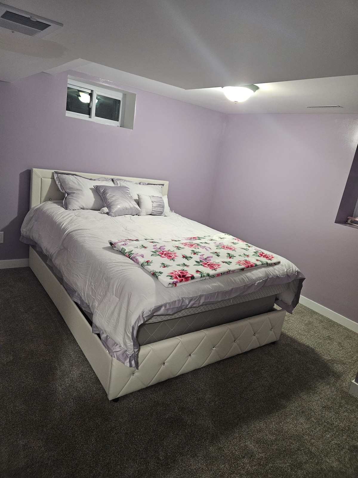 A sleeping area is presented with light purple walls, complemented by plush carpeting. A spacious bed is adorned with soft linens and decorative pillows. Natural light enters through a small window, enhancing the calming ambiance of the room.