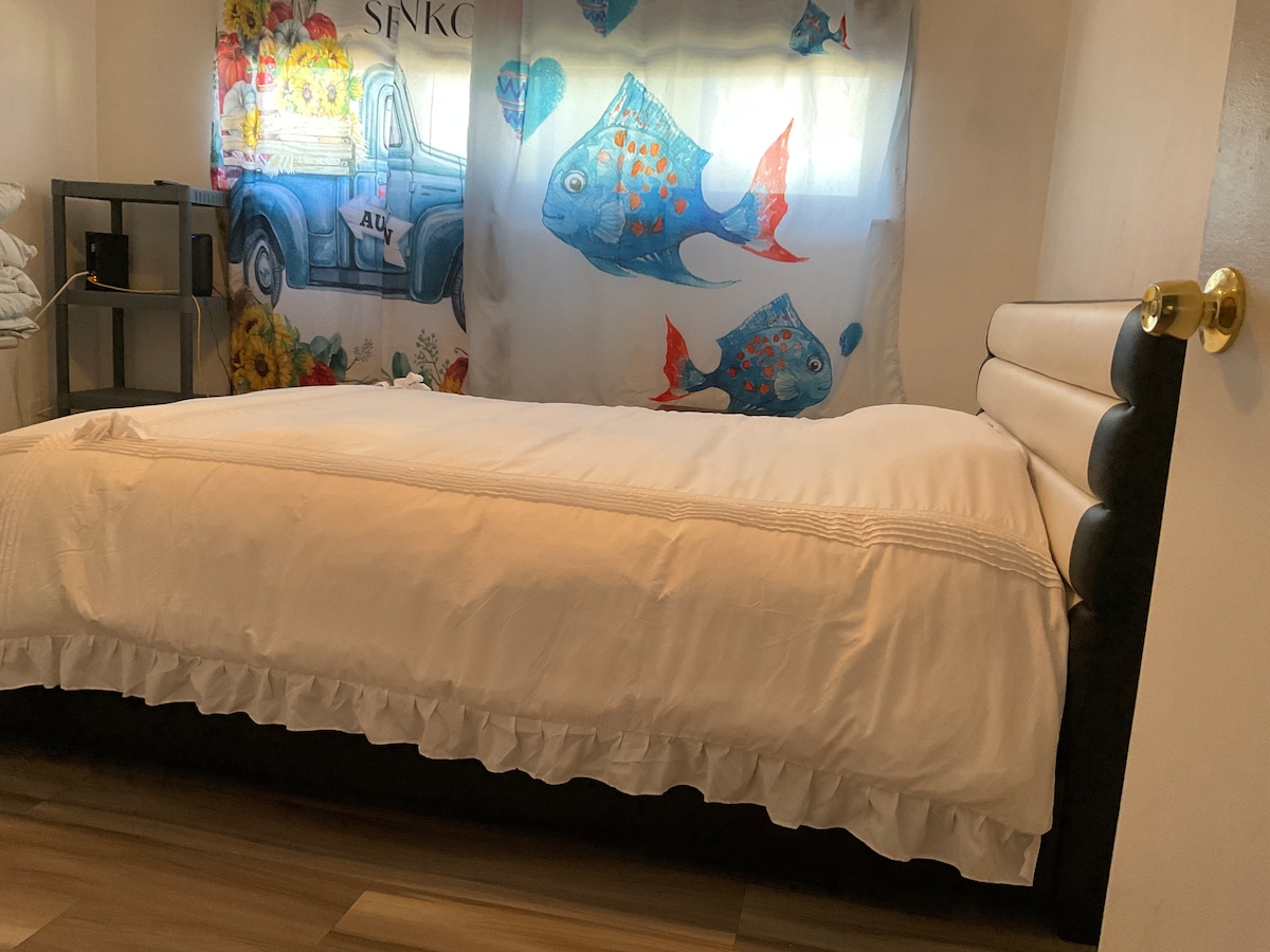A spacious bed with a ruffled white coverlet is positioned against a wall adorned with a colorful fish-themed curtain. A black shelf unit is visible nearby, providing additional storage in the cozy sleeping area.