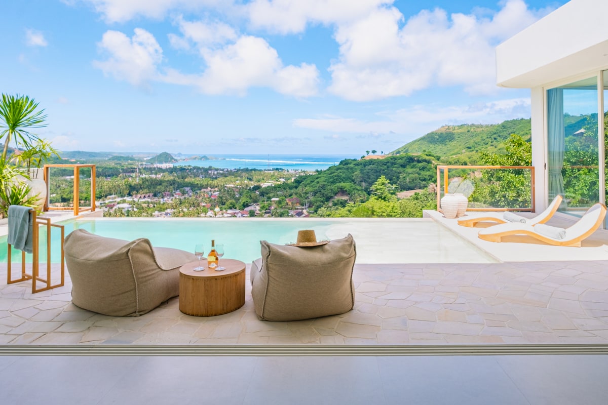 An expansive outdoor seating area features two oversized lounge chairs and a small wooden table. A panoramic view of Kuta Bay is visible beyond the infinity pool, framed by lush greenery and rolling hills under a clear blue sky.