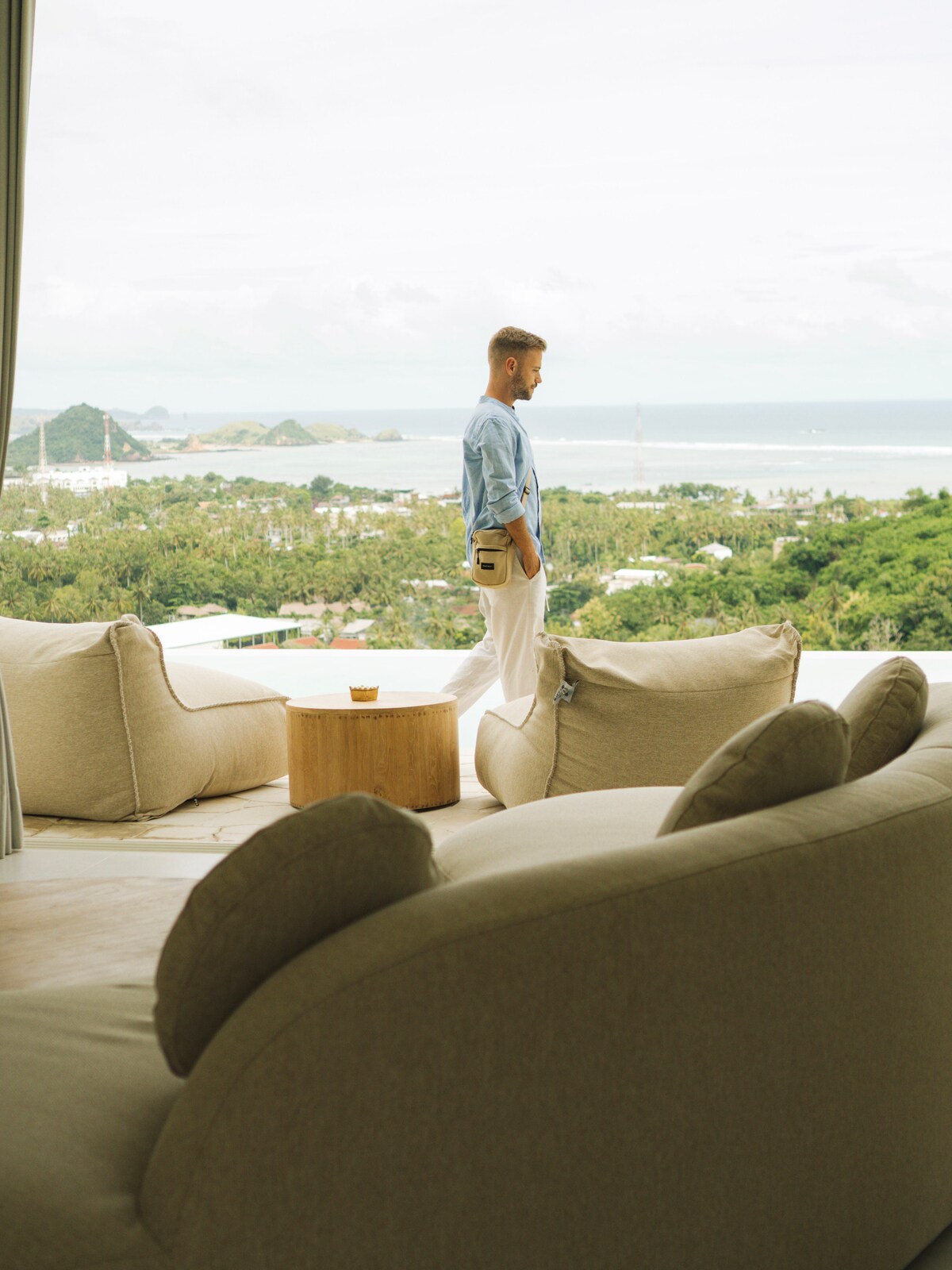 A comfortable seating area features soft, curved furniture with neutral tones. A small wooden table sits nearby, while large windows provide expansive views of the lush landscape and Kuta Bay in the distance. Natural light enhances the serene ambiance of the space.