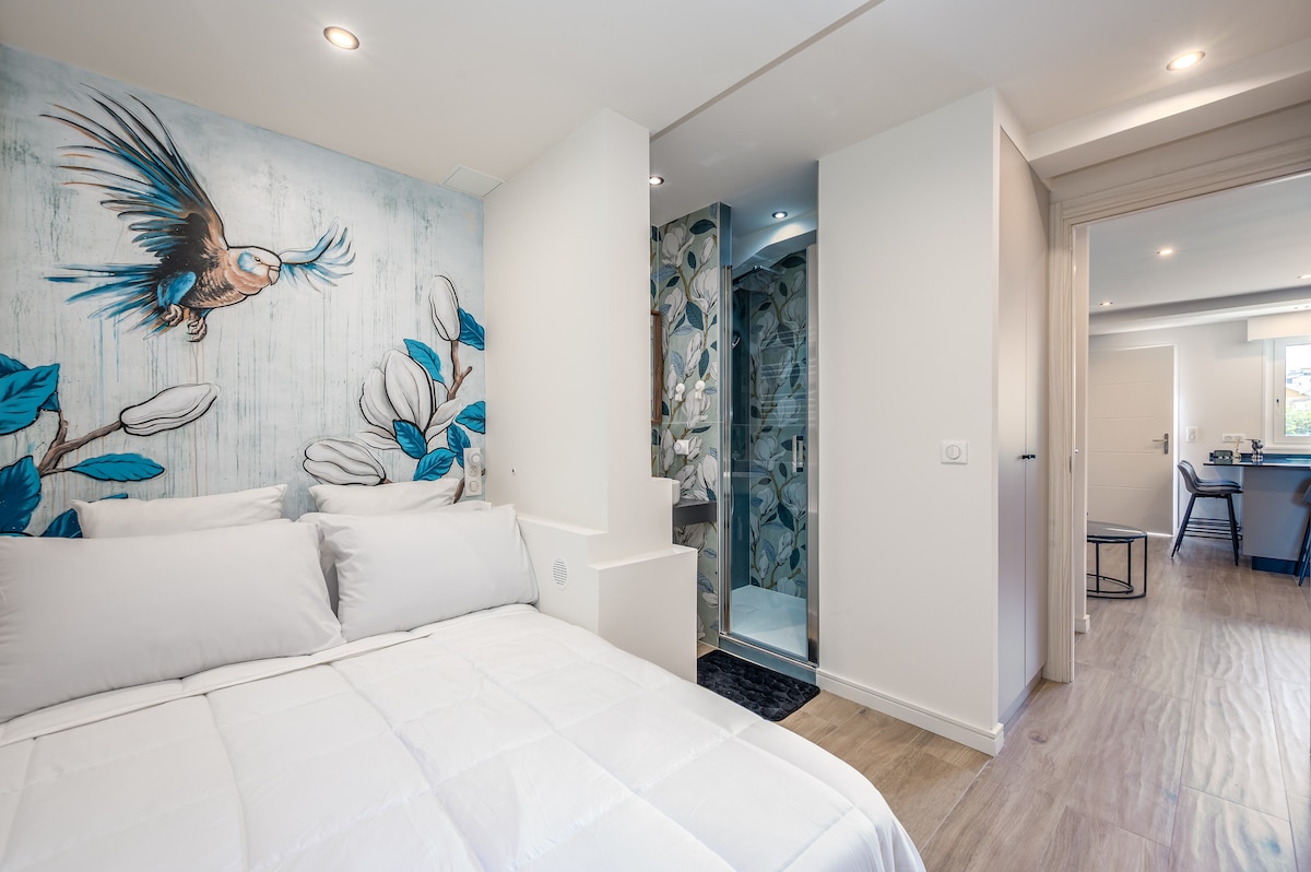 A comfortable bedroom features a spacious bed with crisp white bedding. A vibrant mural of a bird is displayed on one wall. An en-suite bathroom is accessed through a sliding door, showcasing floral tiles and modern fixtures.