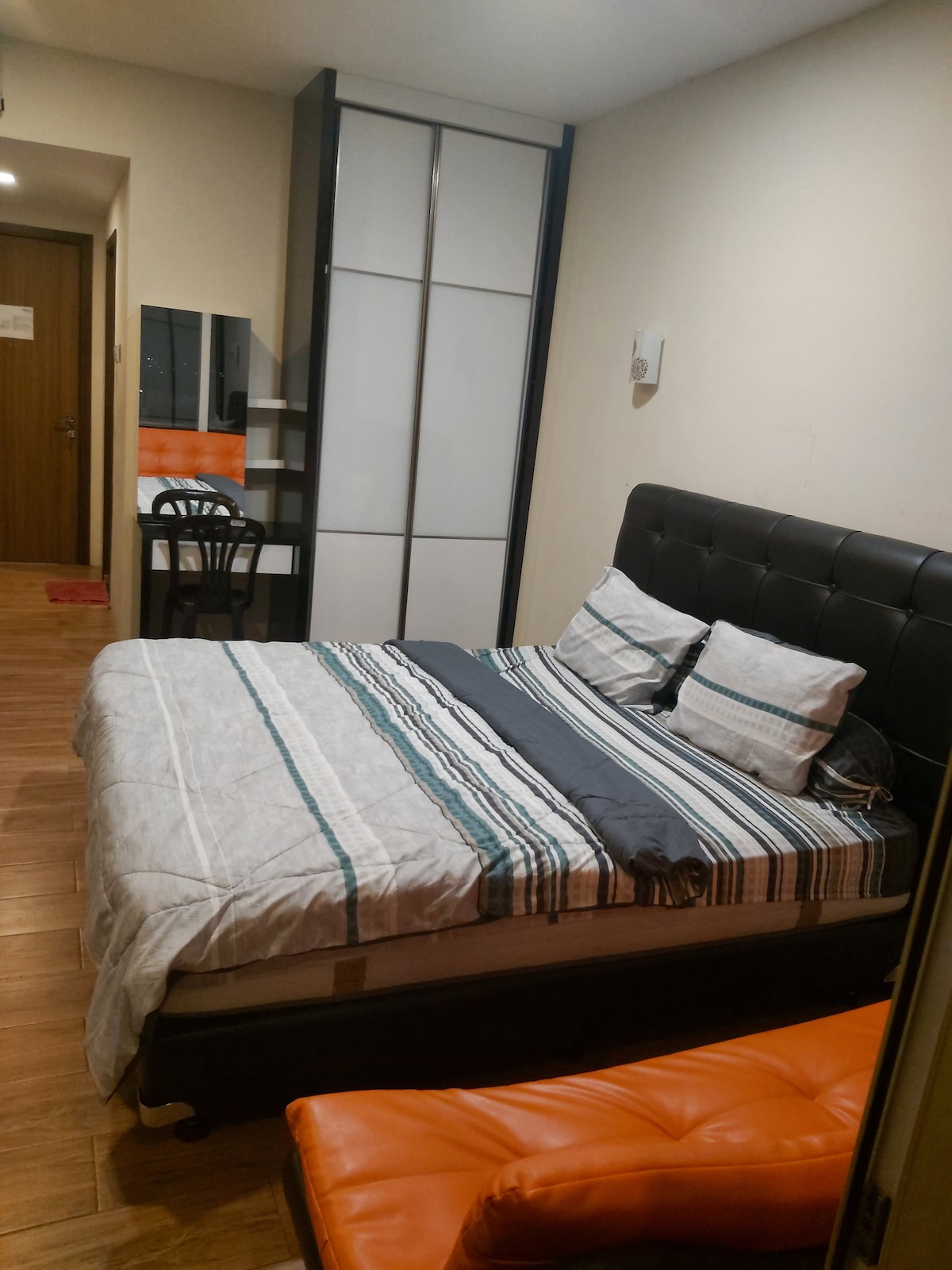 A sleeping area features a dark upholstered bed with striped bedding. A chair sits beside a small desk, and a mirrored wardrobe stands against the wall. Warm wooden flooring adds to the cozy appearance of the room.