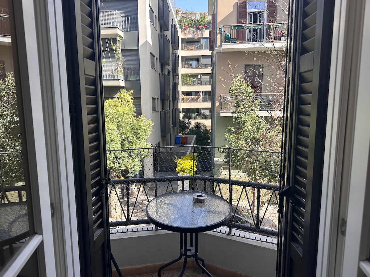 A small balcony is visible, featuring a round table with a cigarette holder at its center. Two sets of shutters are opened wide, providing a view of neighboring buildings and greenery. The surrounding area appears lively, with plants visible in the foreground.