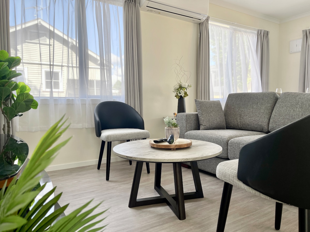 A modern living area features a comfortable gray sofa alongside two chairs—one in a black finish and another in light fabric. A round table sits at the center, adorned with a decorative tray and small floral arrangements. Natural light fills the space through sheer curtains.