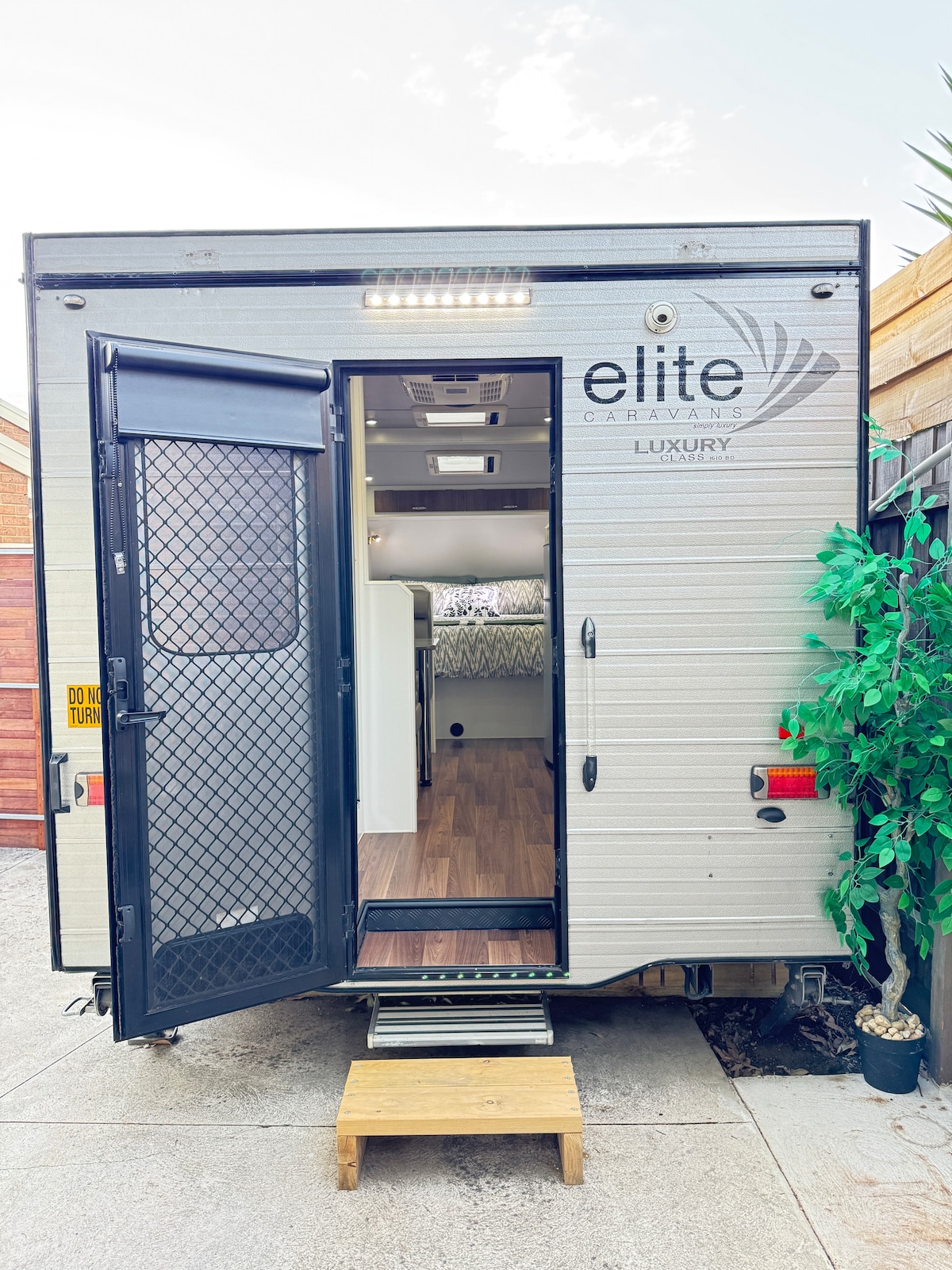 The exterior of a modern caravan is visible, showcasing a double door entrance with a mesh screen. A small wooden step leads up to the open doorway, revealing a glimpse of the interior space. Green foliage is positioned nearby, enhancing the surrounding area.