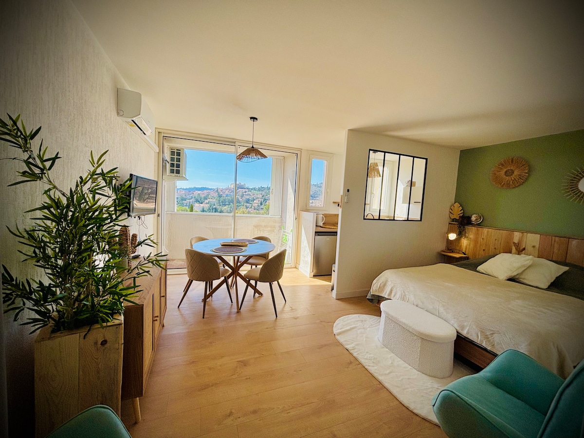 A well-decorated studio features a spacious sleeping area with a large bed and natural light streaming through the window. A dining table is set for four, with stylish chairs nearby. Indoor plants add a touch of greenery, complementing the modern decor.