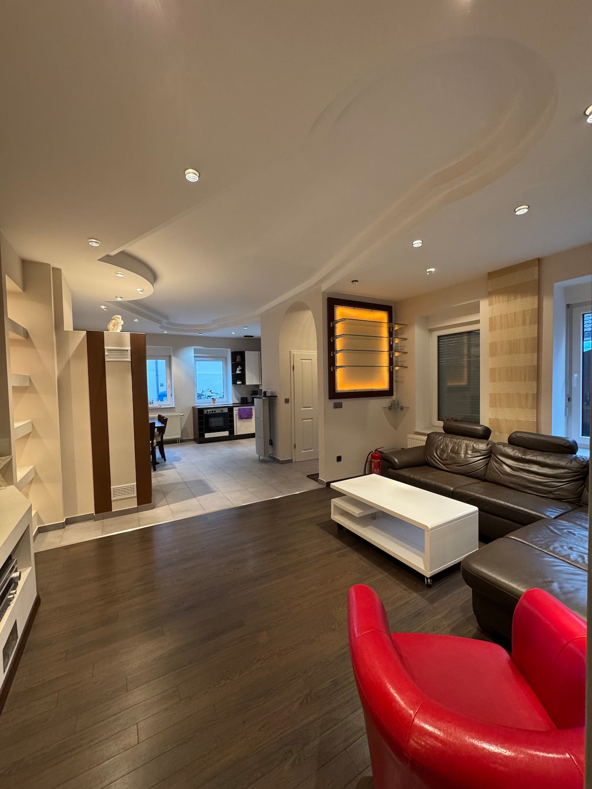 The living area features a modern design with dark hardwood flooring and a mix of bright and neutral tones. A leather sectional sofa offers comfortable seating. A white coffee table sits in the center, while built-in shelves and soft lighting enhance the space's contemporary feel.