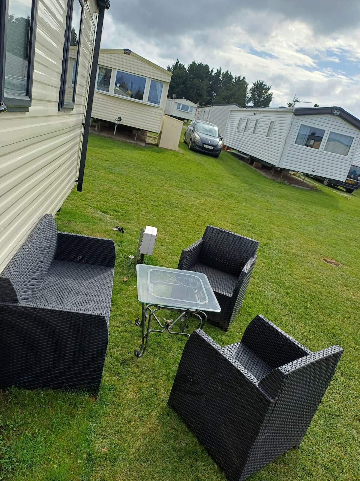 An outdoor seating area features two armchairs and a loveseat arranged around a glass-topped table. Green grass surrounds the seating, providing a comfortable space for relaxation. Nearby, additional caravans are positioned, enhancing the sense of community within the park.