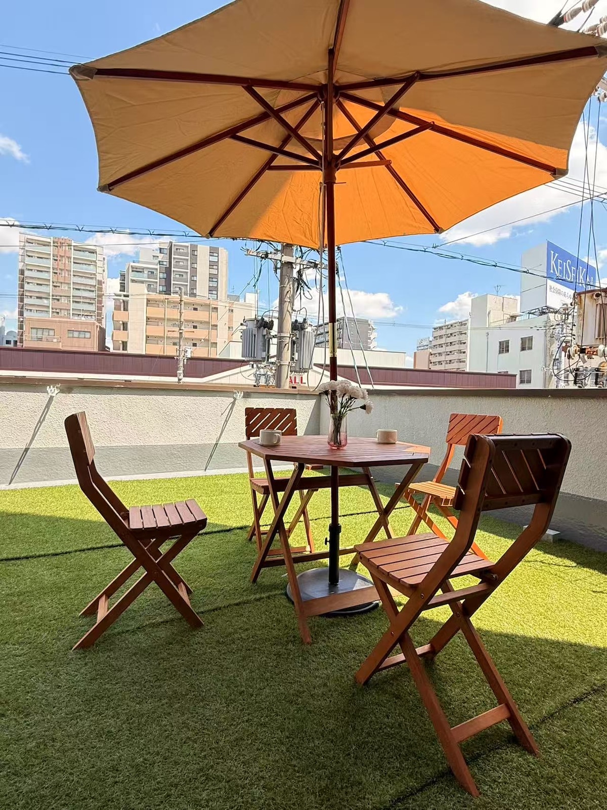 An outdoor terrace features a round table surrounded by four folding chairs. A large umbrella casts shade over the table, providing a comfortable space to relax. The area is adorned with artificial grass, and city buildings can be seen in the background against a clear blue sky.