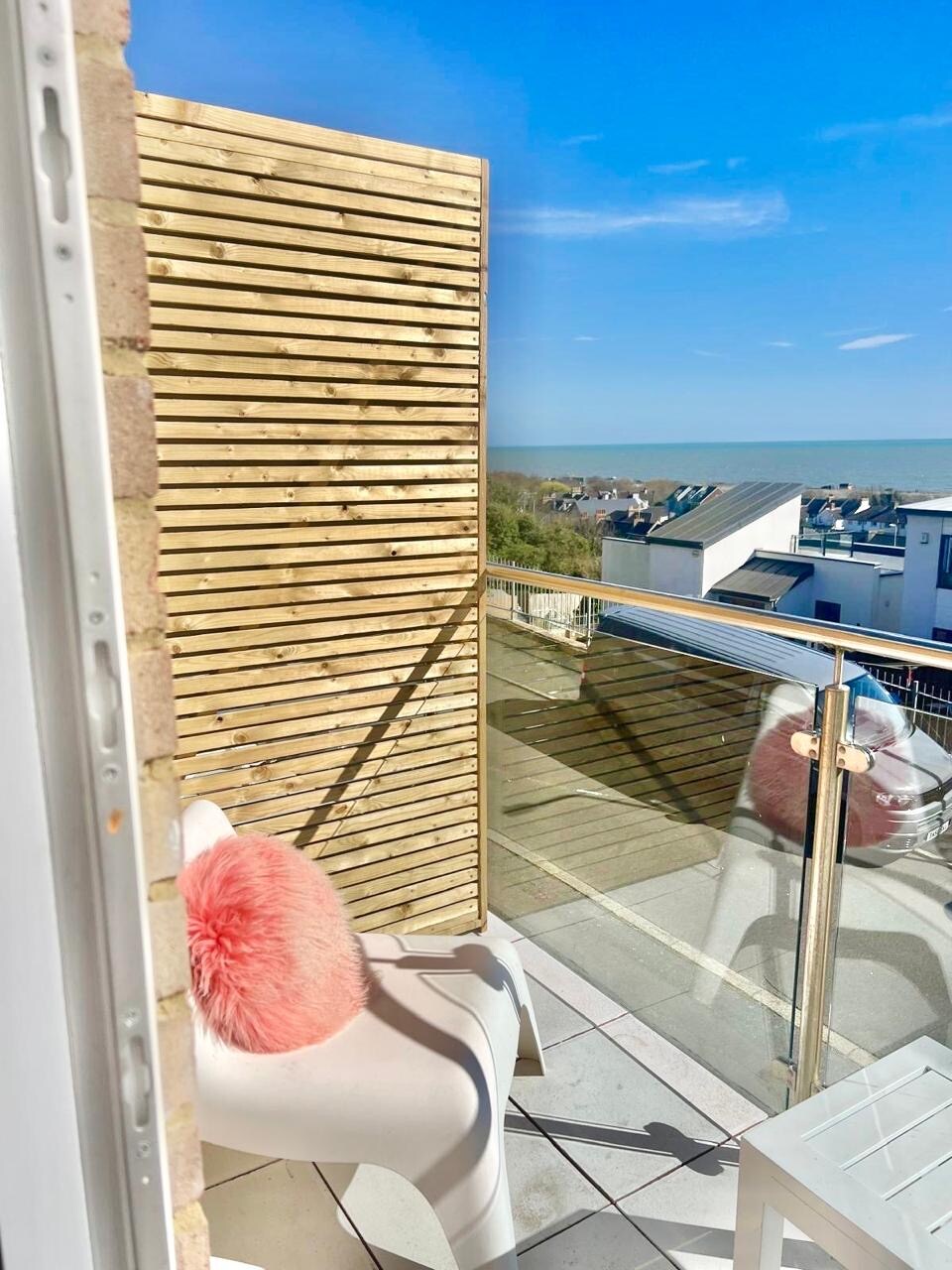 A small balcony features a transparent glass railing, providing a clear view of nearby rooftops and the sea in the distance. A white chair sits in the corner, accented by a soft pink decorative cushion. Sunlight illuminates the space, enhancing the vibrant colors.