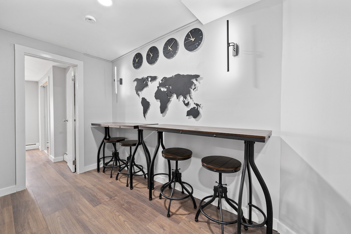 A modern dining area features two high tables accompanied by three circular wooden stools. A world map is displayed on the wall, complemented by four decorative clocks. Soft lighting is provided by sleek wall sconces, creating a functional and stylish space.