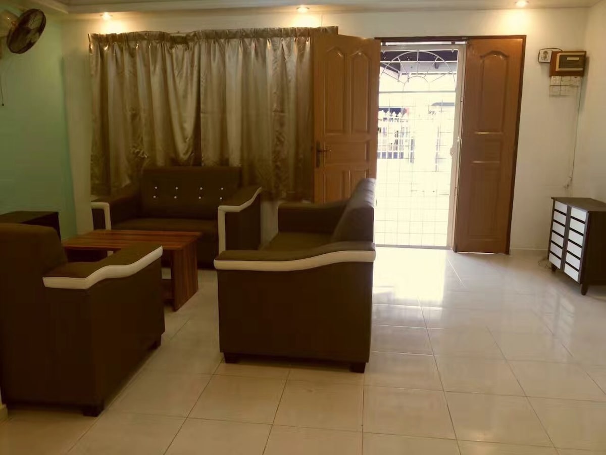 A living area is furnished with a set of brown sofas and a wooden coffee table. Natural light enters through the open door, illuminating the tiled floor. Soft beige curtains frame the window, complementing the light color scheme of the room.